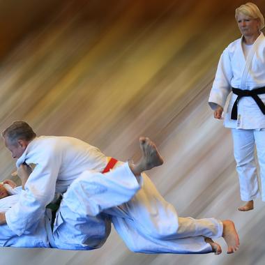 Zwei Judokas in Aktion, einer wirft den anderen auf eine Matte, während eine dritte Person zuschaut.