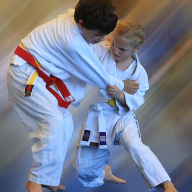 Zwei Kinder im Judoanzug kämpfen, das Mädchen hält den Jungen am Arm, während beide barfuß auf einer Matte stehen.