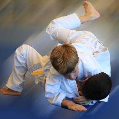 Zwei Kinder im Judogi kämpfen auf einer blauen Matte in einem dynamischen Bodenkampf.