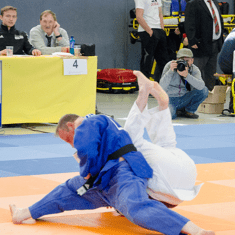 Zwei Judokas kämpfen auf einer Matte, einer in blauer und der andere in weißer Kleidung. Zuschauer im Hintergrund.
