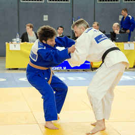 Zwei Judokas kämpfen auf einer Matte; einer trägt einen blauen, der andere einen weißen Judoanzug.