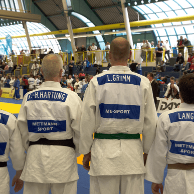 Vier Judokas stehen Rücken an Rücken, in weißen Anzügen mit blauen Namensschildern auf einer Wettkampfmatte.