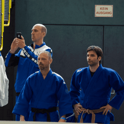 Drei Männer in blauen Judogi stehen am Rand einer Judohalle, während einer ein Smartphone hält. Im Hintergrund ein Ausgangsschild.