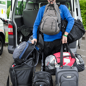 Junger Mann trägt mehrere Reisetaschen und Rucksäcke, steht vor einem geöffneten Transporter.