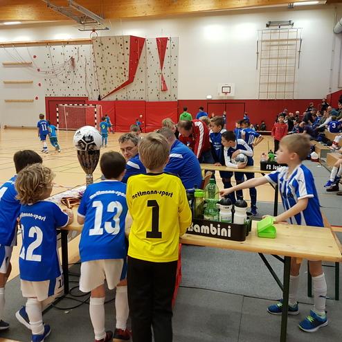 Junge Fußballspieler in blauen Trikots stehen an einem Tisch mit Pokal und Getränken in einer Sporthalle.