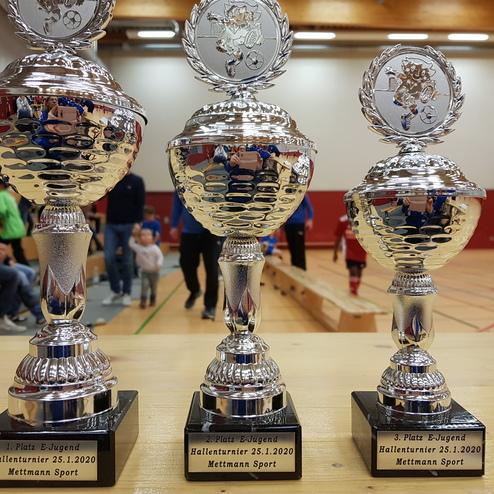 Drei silberne Pokale für den 1., 2. und 3. Platz eines Hallenturniers, mit Inschriften auf schwarzem Sockel.