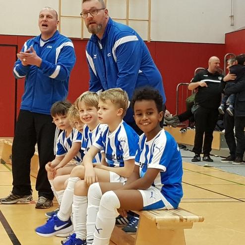 Trainer steht hinter einer Gruppe von fünf lächelnden Jungen in Fußballtrikots, während Zuschauer im Hintergrund stehen.