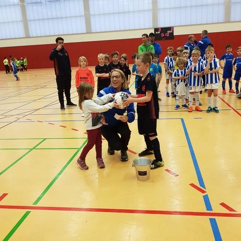 Ein Mädchen übergibt einem Trainer einen Fußball, während Kinder in Sportkleidung auf einem Hallenboden warten.