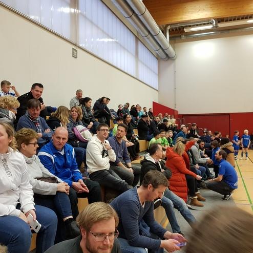 Zuschauer sitzen in einer Sporthalle auf Bänken, während ein Kindersportspiel im Hintergrund stattfindet.