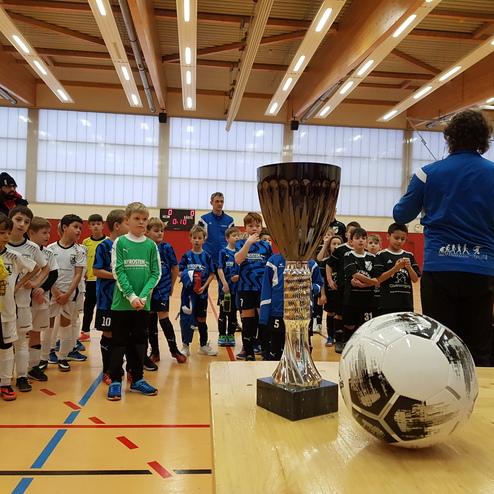 Versammlung von Kindern in Fußballtrikots vor einem Pokal und einem Fußball, während ein Trainer spricht.