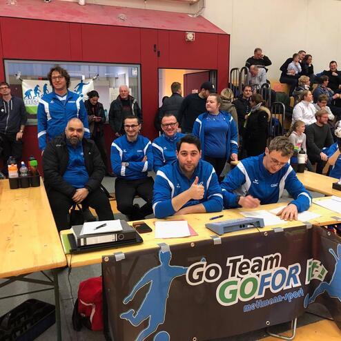 Gruppe von Coaches in blauen Jacken sitzt an einem Tisch, neben einer Trophäe und Getränken in einer Sporthalle.