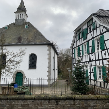 Zwei historische Gebäude: eine weiße Kirche mit spitzem Dach und ein Fachwerkhaus mit grünen Fensterläden.