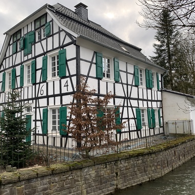 Fachwerkhaus mit grünem Fensterladen, umgeben von Bäumen und einem Wasserlauf, Sicht von der Seite.