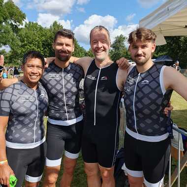 Vier Sportler in enger Triathlonbekleidung posieren im Freien, umgeben von Grünflächen und einem blauen Himmel.