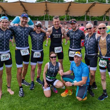 Gruppe von acht Triathleten in Sportoutfits, auf einer Wiese in Sportarena, vor Wettkampfbeginn.