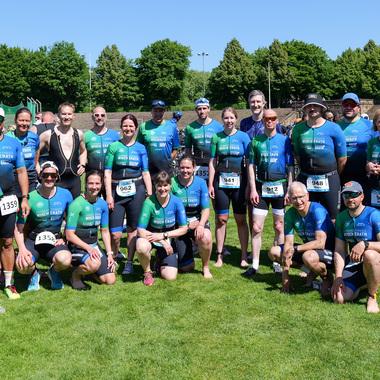 Gruppenselfie von Athleten in Sportanzügen auf einer Wiese, umgeben von Bäumen und Zuschauern im Hintergrund.