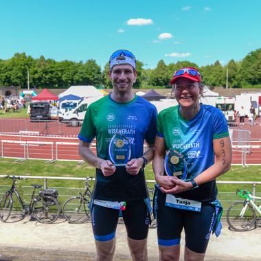 Zwei Athleten mit Medaillen stehen lächelnd auf einer Sportveranstaltung, mit Zuschauerbereichen und Radfahrern im Hintergrund.