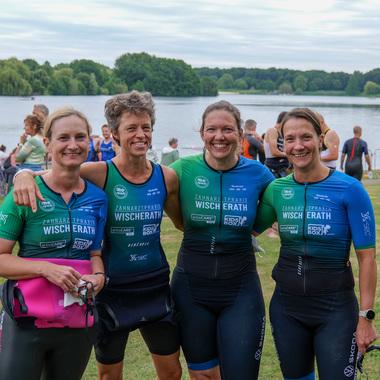 Vier Frauen in Sportanzügen lächeln am Ufer eines Sees, im Hintergrund eine Gruppe von Zuschauern und Teilnehmern.