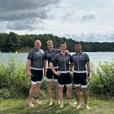Vier Männer in Sportbekleidung stehen am Ufer eines Gewässers, umgeben von Bäumen und einem bewölkten Himmel.