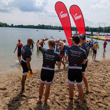Gruppierung von Schwimmern in Wettkampfanzügen am Sandstrand eines Sees, umgeben von Booten und Flaggen.