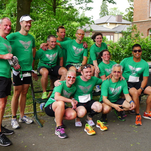 Gruppe von zwölf Personen in grünen T-Shirts lächelt auf einer Bank, umgeben von Bäumen und einem Schulgebäude.