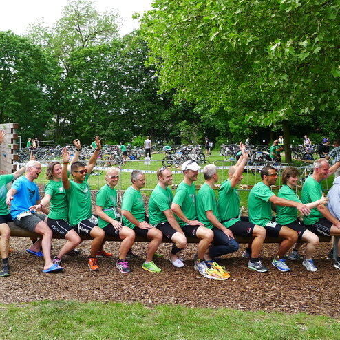 Gruppe von 15 Personen in grünen Shirts, die auf einer Bank sitzen und den Arm heben, um zum Fotoshooting zu posieren.