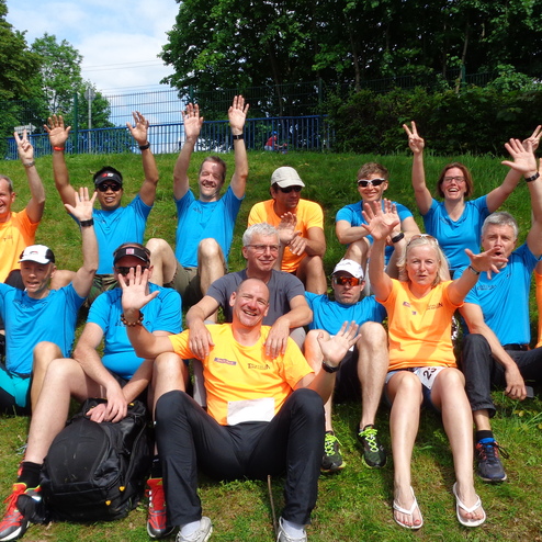 Gruppenshot von 16 Menschen in bunten T-Shirts, die auf einer Wiese sitzen und lächeln, während sie die Hände heben.