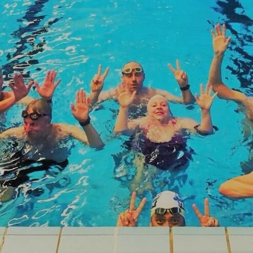 Gruppenselfie von Schwimmern im Wasser, die freundlich in die Kamera winken und zwei Finger hochhalten.