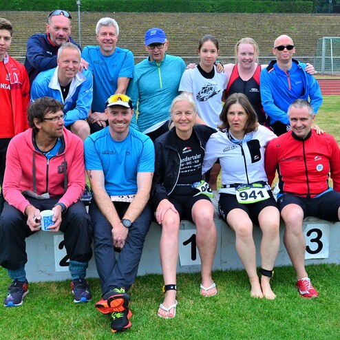 Gruppenshot von 12 Sportlern in Sportkleidung, sitzend und stehend auf einer Wiese vor einem Sportplatz.