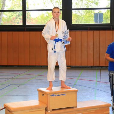 Junge in Judogi mit Goldmedaille und Urkunde steht auf dem Siegertreppchen, daneben ein Mann mit Unterlagen.
