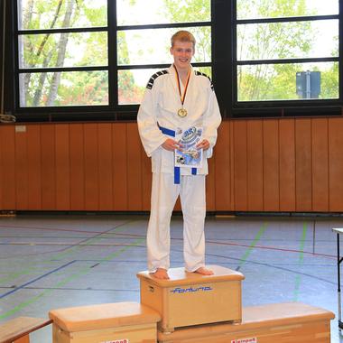 Junger Sportler im Judogi steht auf dem Siegerpodest, hält Medaille und Urkunde, neben ihm ein Mann mit Pokalen.