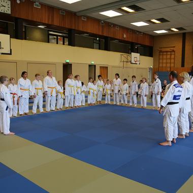Versammlung junger Kampfsportler in weißen Anzügen auf einer blauen Matte in einer Sporthalle.