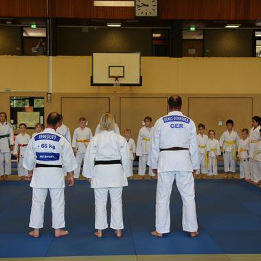 Judo-Training mit mehreren Teilnehmern in weißen und blauen Judogis, die auf einer Matte in einer Sporthalle versammelt sind.