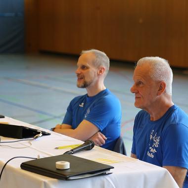 Zwei Männer in blauen T-Shirts sitzen an einem Tisch mit Schreibmaterial in einer Sporthalle.