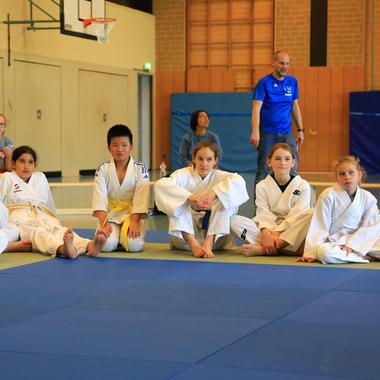 Gruppe von Kindern in weißem Judogi sitzt auf einer Judomatte, während Erwachsene im Hintergrund stehen und zuschauen.