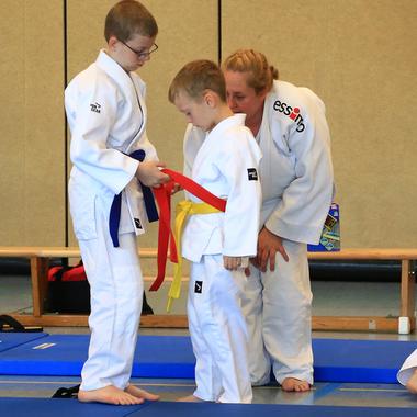 Zwei Kinder im judogi binden einem Jungen mit gelbem Gürtel einen roten Gürtel an, eine Trainerin beobachtet.