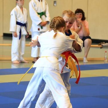 Zwei Kinder im Judoanzug kämpfen auf einer blauen Matte, während Zuschauer im Hintergrund beobachten.