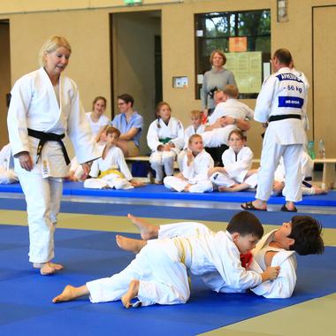 Zwei Kinder im Judoanzug üben eine Technik auf einer blauen Judomatte, während eine Trainerin zuschaut.