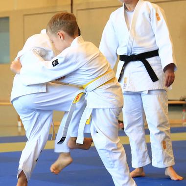 Zwei Kinder in Judogis üben eine Wurftechnik auf einer blauen Judomatte, während eine Trainerin zuschaut.