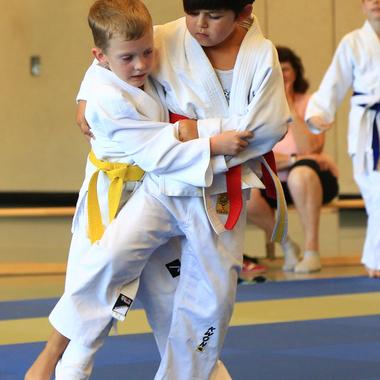Zwei Kinder im Judogi üben gegenseitig eine Technik auf einer blauen Judomatte, Zuschauer im Hintergrund.