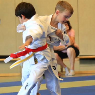 Zwei Kinder in weißen Judoanzügen kämpfen auf blauen Matten, eines mit rotem Gürtel, das andere mit gelbem Gürtel.