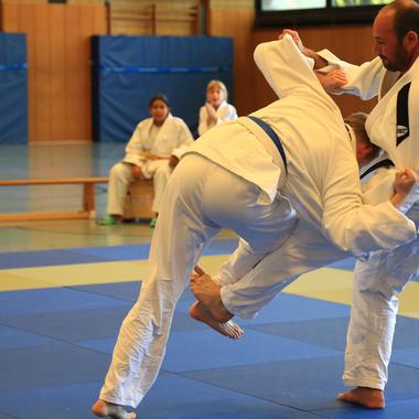 Zwei Judoka im Training, einer führt einen Wurf aus, während andere Zuschauer auf Matten sitzen.