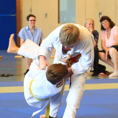 Zwei Kinder im Judoanzug kämpfen auf einer blauen Matte, Zuschauer sitzen im Hintergrund.