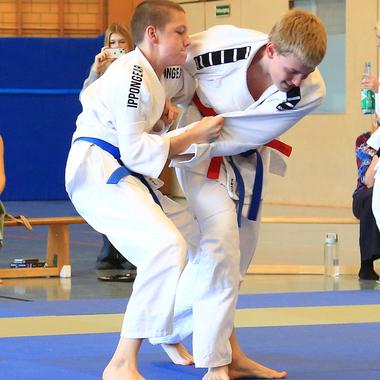 Zwei Judokas in weißem Gi kämpfen auf einer blauen Matte, einer greift den anderen an. Zuschauer im Hintergrund.