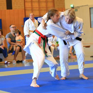 Zwei Judokas kämpfen auf einer blauen Matte, Zuschauer beobachten das Geschehen im Hintergrund.