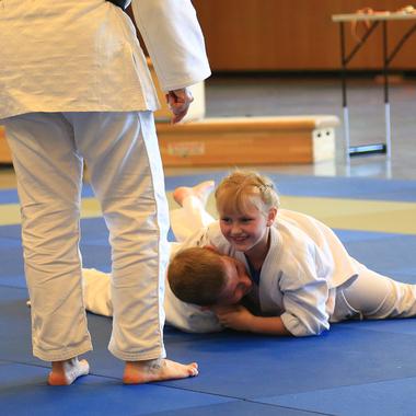 Zwei Kinder im weißen Judogi ringen auf einer blauen Judomatte, ein Erwachsener beobachtet die Szene.