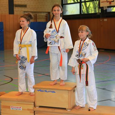 Drei Mädchen in weißen Karate-Anzügen stehen auf dem Siegertreppchen und halten Medaillen und Urkunden.