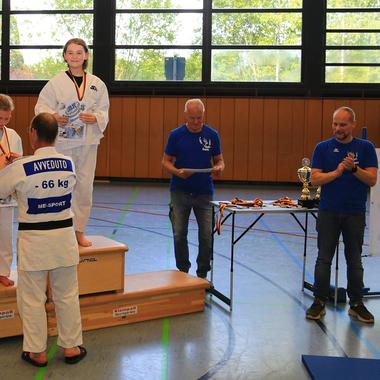 Zwei Judokas stehen auf dem Siegerpodest, die Siegerin trägt eine Medaille, weitere Personen applaudieren im Hintergrund.