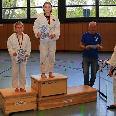 Zwei junge Judokas stehen auf dem Siegertreppchen mit Medaillen und Urkunden, während Trainer und Zuschauer zuschauen.