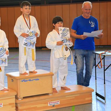 Vier Kinder in Judogi stehen auf einem Treppchen und halten Medaillen sowie Urkunden während einer Auszeichnungszeremonie.